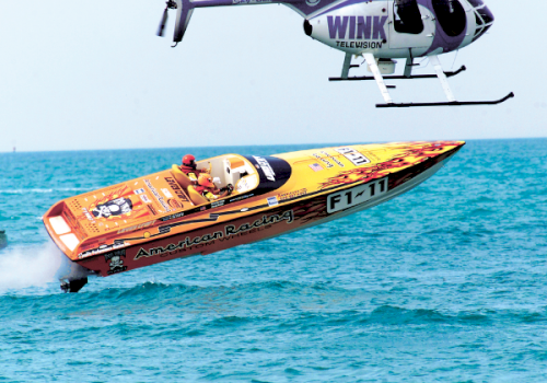 A fast racing speedboat skims across the ocean while a helicopter hovers above it.