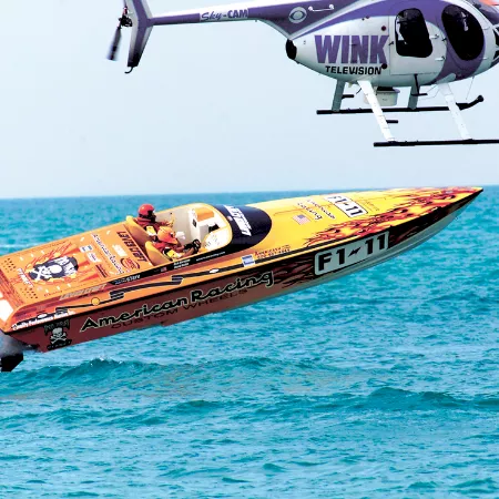 A fast racing speedboat skims across the ocean while a helicopter hovers above it.