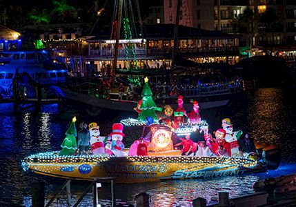 A colorful holiday parade boat with glowing characters floats on a night harbor.