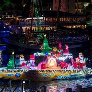 A colorful holiday parade boat with glowing characters floats on a night harbor.