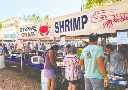 An outdoor seafood stand selling shrimp and crab, with big signs and people browsing tables on a sunny day.