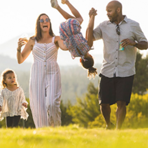 A family of four is enjoying a sunny day outdoors. Two adults swing a child, while another child walks beside them, all smiling.