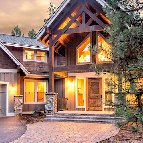 A modern, two-story house with large windows, a gabled roof, and a garage, surrounded by pine trees and illuminated warmly at dusk.