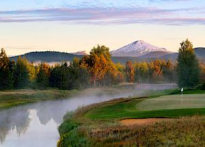 A scenic golf course by a river, surrounded by trees with a distant snow-capped mountain under a partly cloudy sky, bathed in warm sunlight.