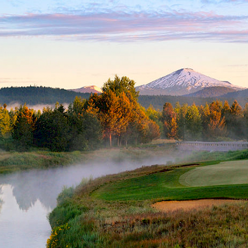 A scenic golf course by a river, surrounded by trees with a distant snow-capped mountain under a partly cloudy sky, bathed in warm sunlight.