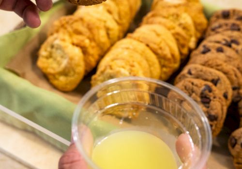 A hand holds a chocolate chip cookie over a tray of assorted cookies; a cup of yellow juice is in the foreground.