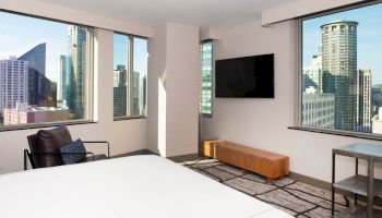 A modern hotel room with floor-to-ceiling windows, city views, a flat-screen TV, a bed, an armchair, and a small table with two stools.