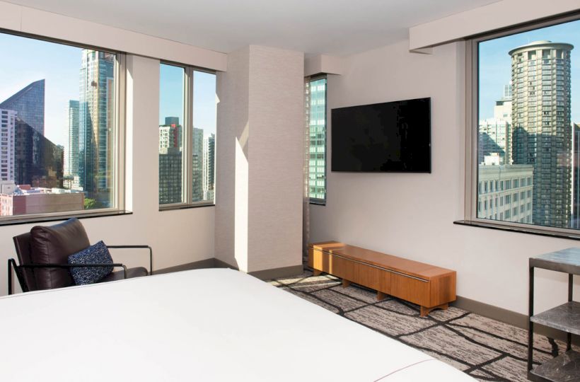 A modern hotel room with floor-to-ceiling windows, city views, a flat-screen TV, a bed, an armchair, and a small table with two stools.