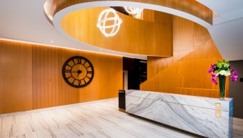 A modern lobby area features a spiral wooden staircase, a marble reception desk, a large wall clock, and a bouquet of colorful flowers.