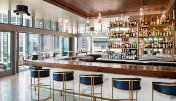The image shows a stylish bar with blue and gold stools, a wooden counter, and shelves filled with bottles. Large windows provide a city view.