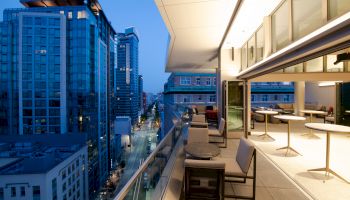 The image shows a cityscape at dusk with modern high-rise buildings and a balcony featuring tables and chairs, overlooking a lit street.