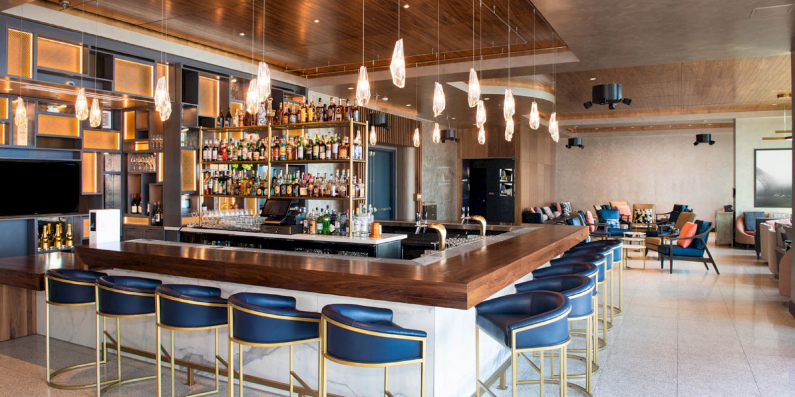 A modern bar with a wooden counter, blue cushioned bar stools, and a well-stocked array of liquors behind. The space features stylish lighting.