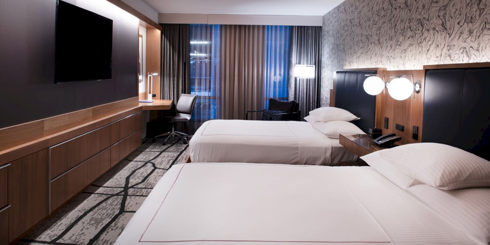 A modern hotel room with two neatly made beds, a wall-mounted TV, a work desk and chair, stylish lighting, and large windows with curtains.