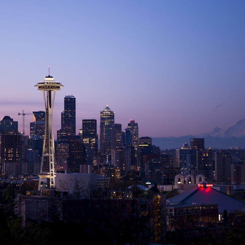 The image shows a dusk cityscape featuring the Seattle skyline with the Space Needle and Mount Rainier in the background, under a twilight sky.