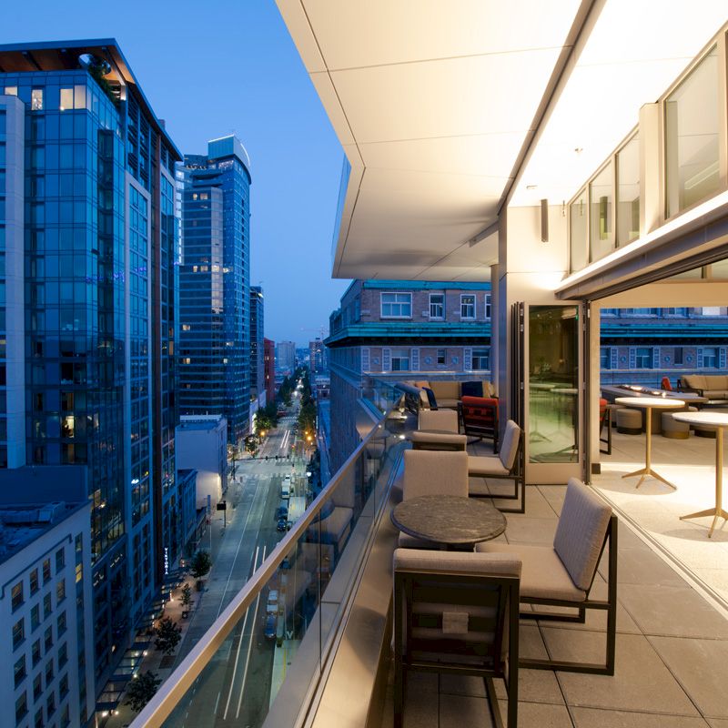 A modern cityscape at twilight with a view from a balcony. The balcony has tables and chairs, overlooking tall buildings and a street below.
