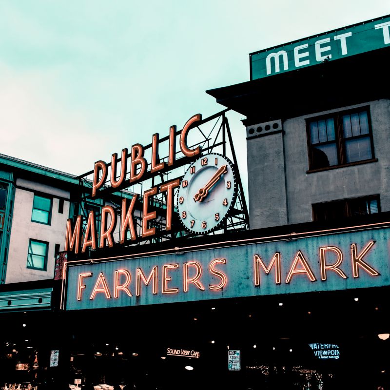 A vibrant neon sign reading "Public Market Farmers Market" sits atop a building under a cloudy sky, with "Meet the" partially visible on an adjacent structure.