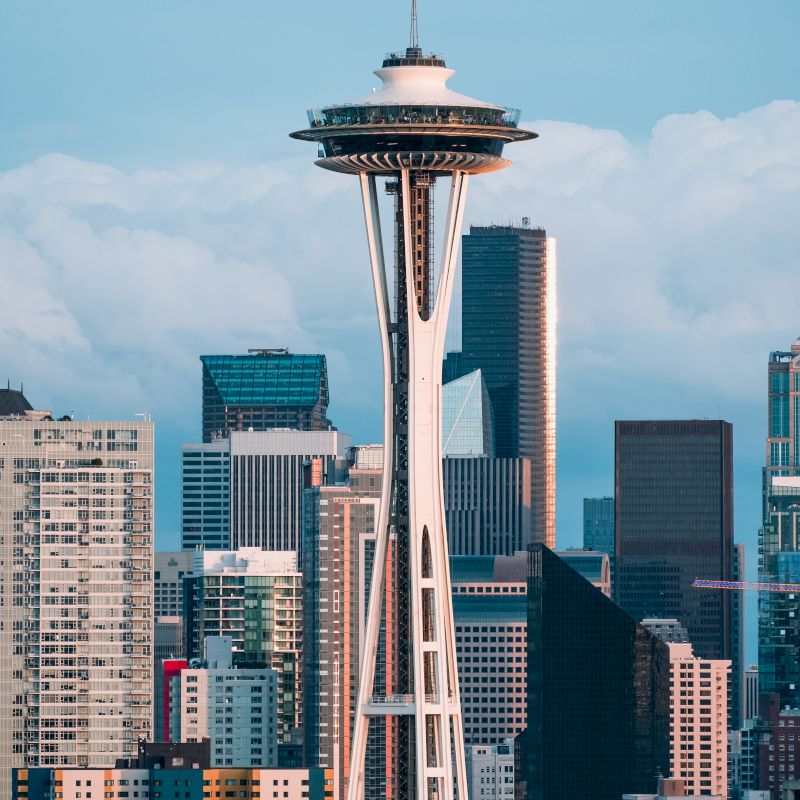 The image shows a cityscape with the iconic Space Needle tower in the foreground, surrounded by various modern buildings and skyscrapers.