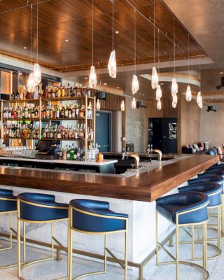 A modern bar with blue barstools, a variety of liquor bottles on shelves, unique hanging lights, and a cozy seating area in the background.