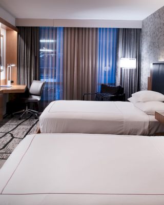 The image shows a modern hotel room with two neatly made beds, a wall-mounted TV, a desk with a chair, and contemporary decor.