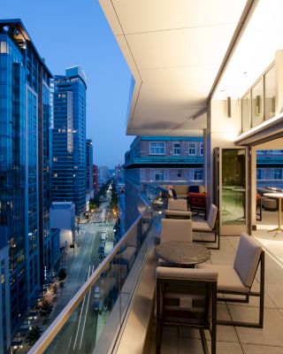 A modern outdoor terrace at dusk with tables and chairs overlooking a city street lined with tall buildings.