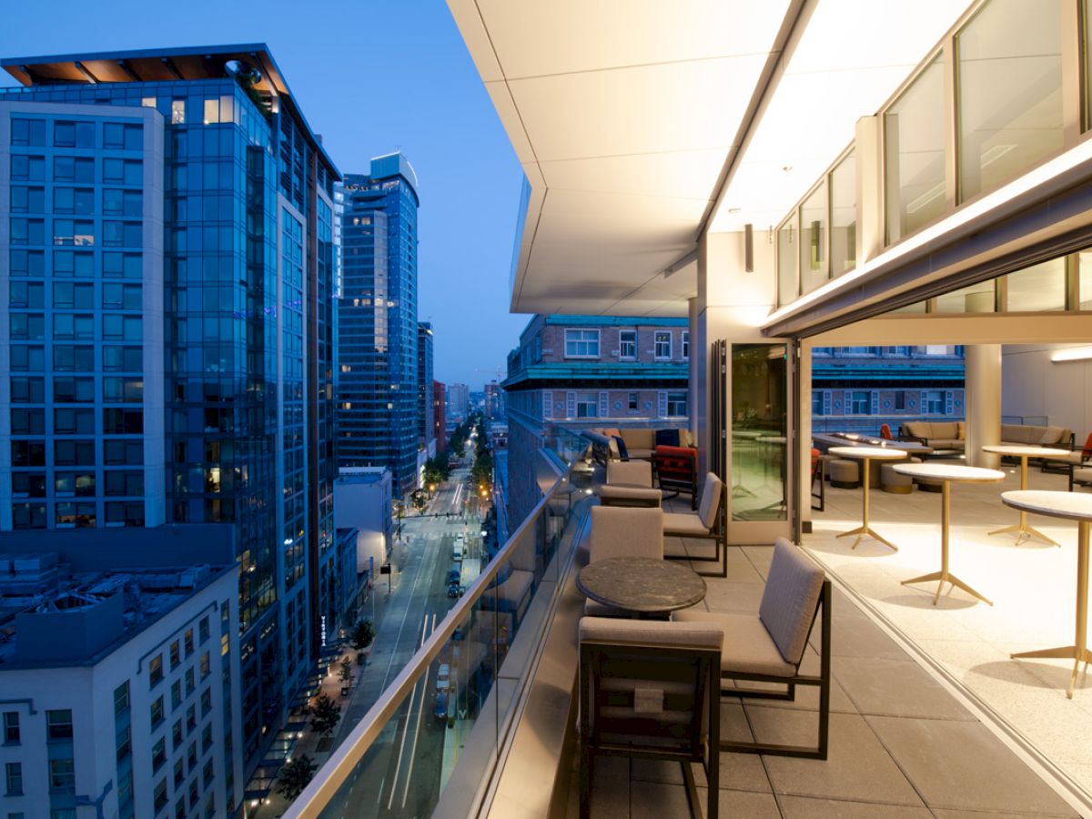 An elevated city view showcases modern high-rise buildings with a furnished rooftop patio, including tables, chairs, and warm lighting at dusk.
