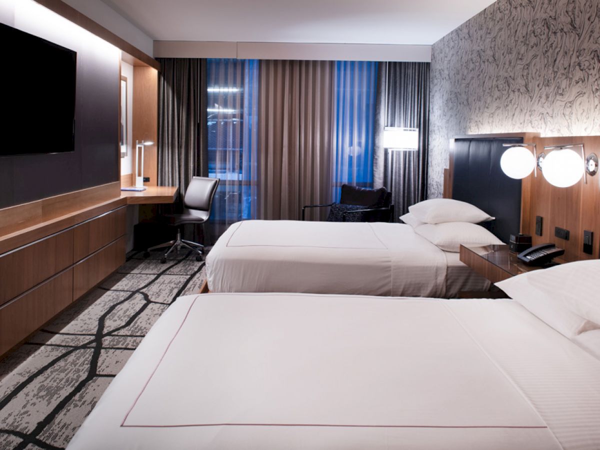 The image shows a modern hotel room with two neatly made beds, a large TV, a work desk with a chair, and floor-to-ceiling curtains.