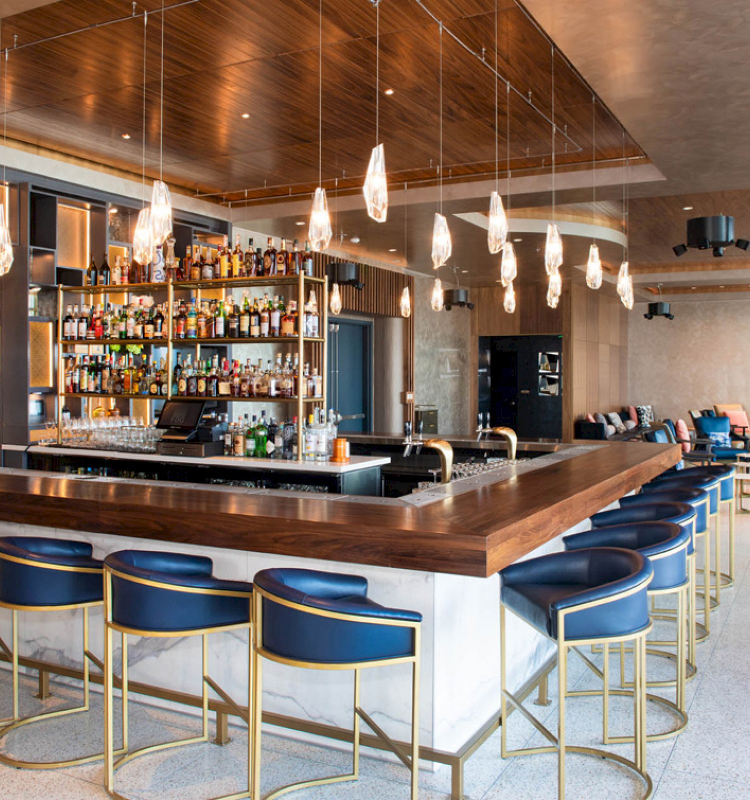 The image shows a stylish bar with modern lighting, a well-stocked liquor shelf, blue barstools with gold accents, and a cozy seating area in the background.
