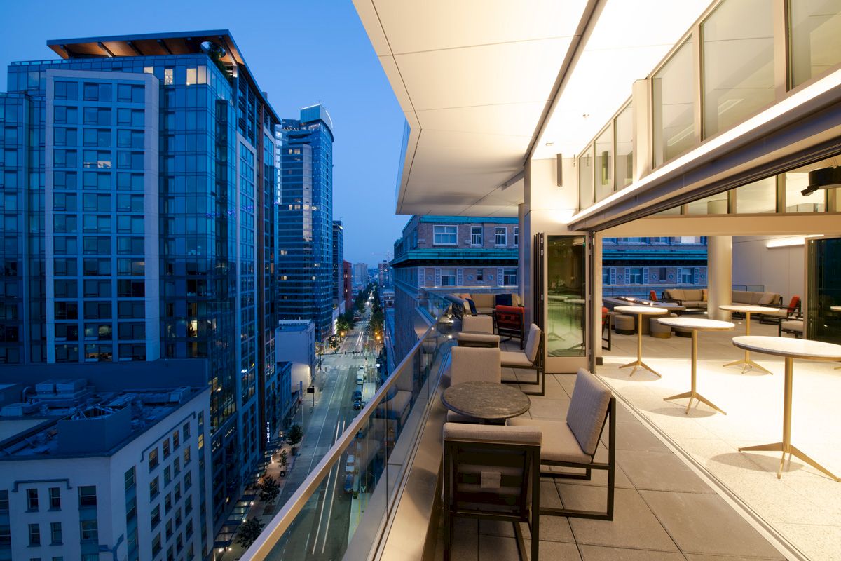 A modern outdoor patio area with tables and chairs, with a view of a cityscape featuring tall buildings at dusk.