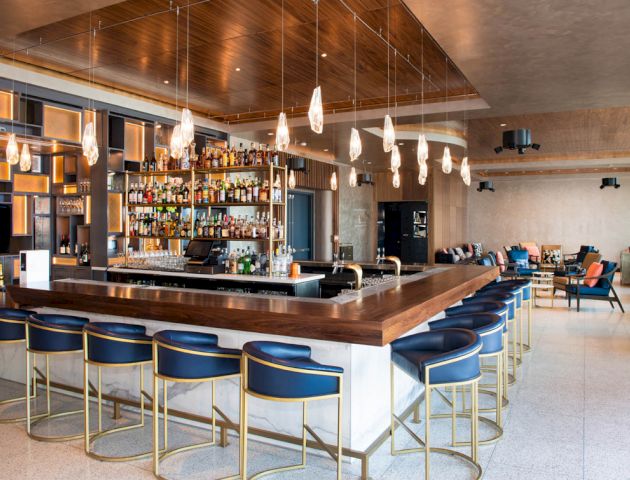 This image shows a modern bar with a large counter, bar stools, hanging lights, and a well-stocked shelf of bottles behind the counter.