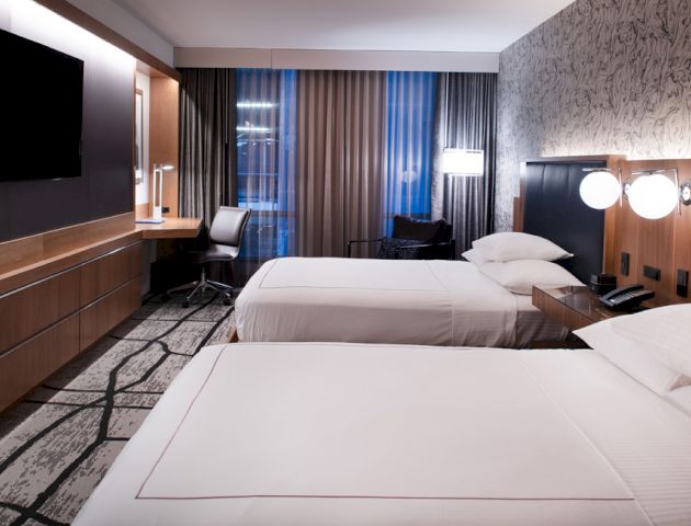 The image shows a modern hotel room with two double beds, a desk and chair, a large TV on the wall, and stylish decor and lighting.