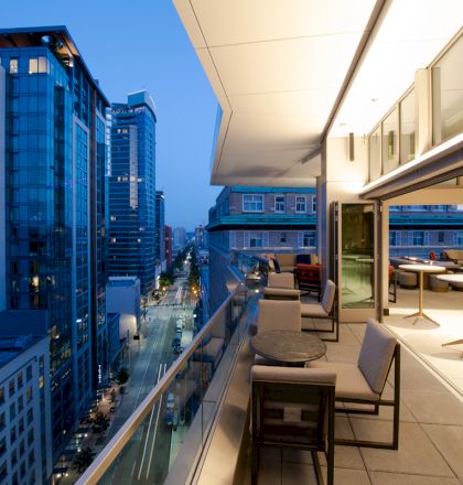 This image shows a modern cityscape at dusk viewed from a balcony with outdoor furniture and glass railings, overlooking tall buildings and a street.