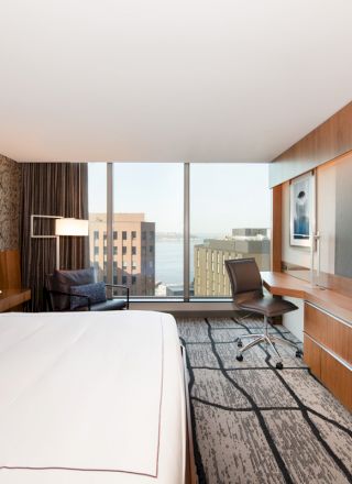 A modern hotel room with a large bed, wall-mounted TV, desk, chair, and floor-to-ceiling windows with a city view.