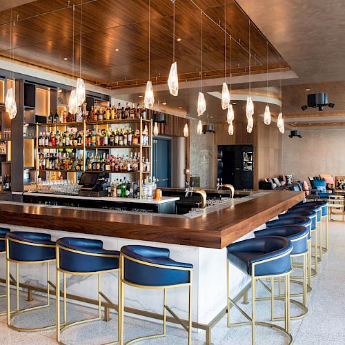 This image shows a modern bar with high chairs, a fully stocked bar, and hanging lights in a stylish and spacious setting.