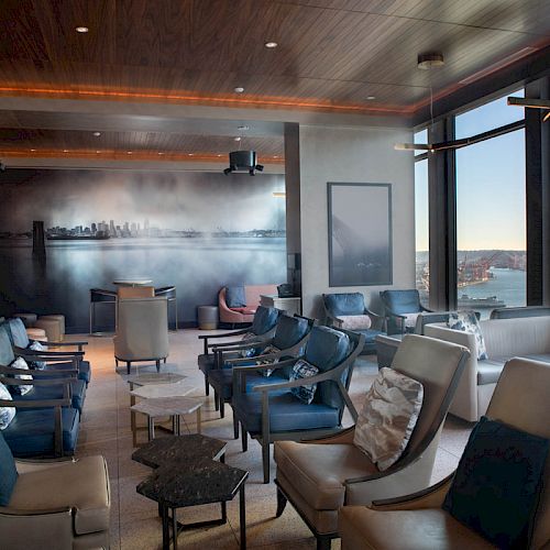 The image shows a modern lounge area with multiple armchairs, tables, and large windows offering a scenic view of a waterfront and ferris wheel.