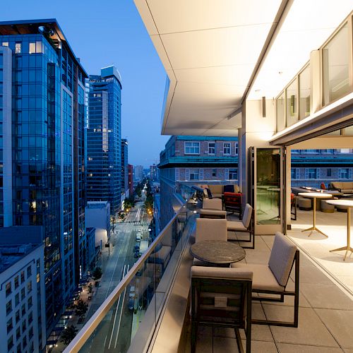A modern cityscape view from a glass-walled balcony at dusk, featuring tables, chairs, and a partially open indoor seating area.