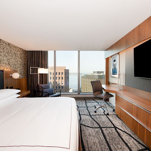 A modern hotel room features a king-size bed, a large flat-screen TV, a work desk, and a city view through wide windows, creating a comfortable stay.