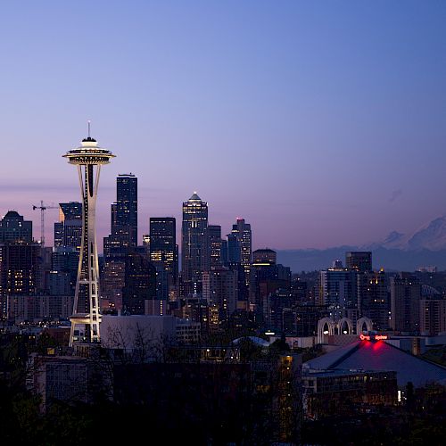 A twilight view of Seattle’s skyline featuring the Space Needle, with the Mount Rainier in the background, and city buildings lit with evening lights.