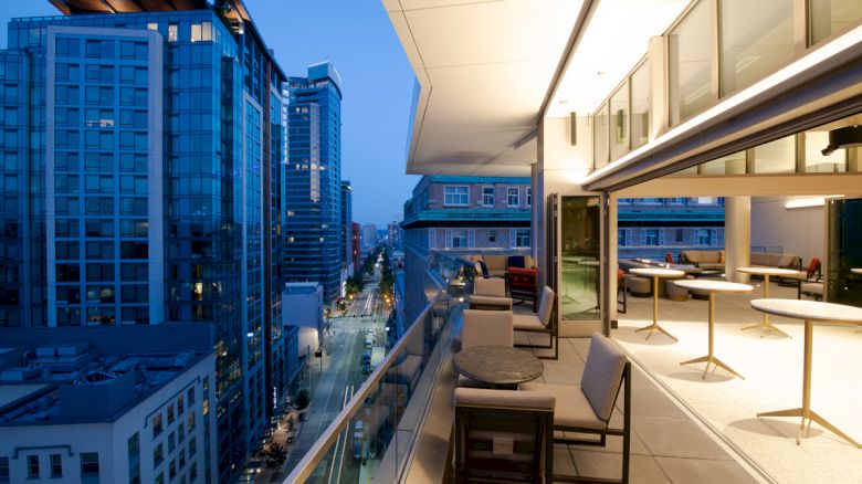 A modern balcony with tables and chairs overlooking a cityscape with tall buildings during the evening, with lights illuminating the area.
