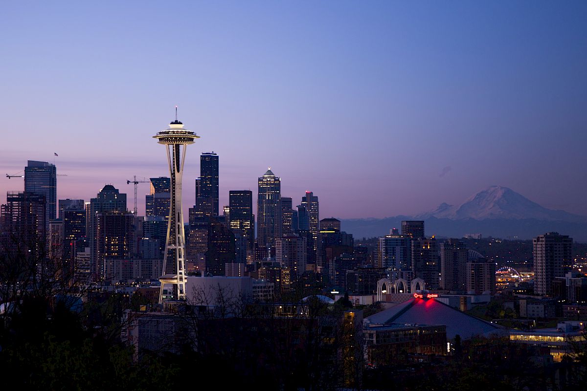 A twilight view of Seattle's skyline with the Space Needle prominently in the center, featuring nearby buildings and a mountain in the background.