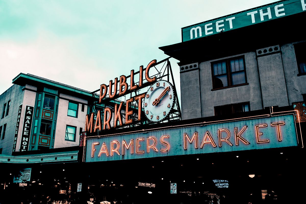The image shows a public market building with neon signs reading "Public Market" and "Farmers Market." A clock is also visible in the center.
