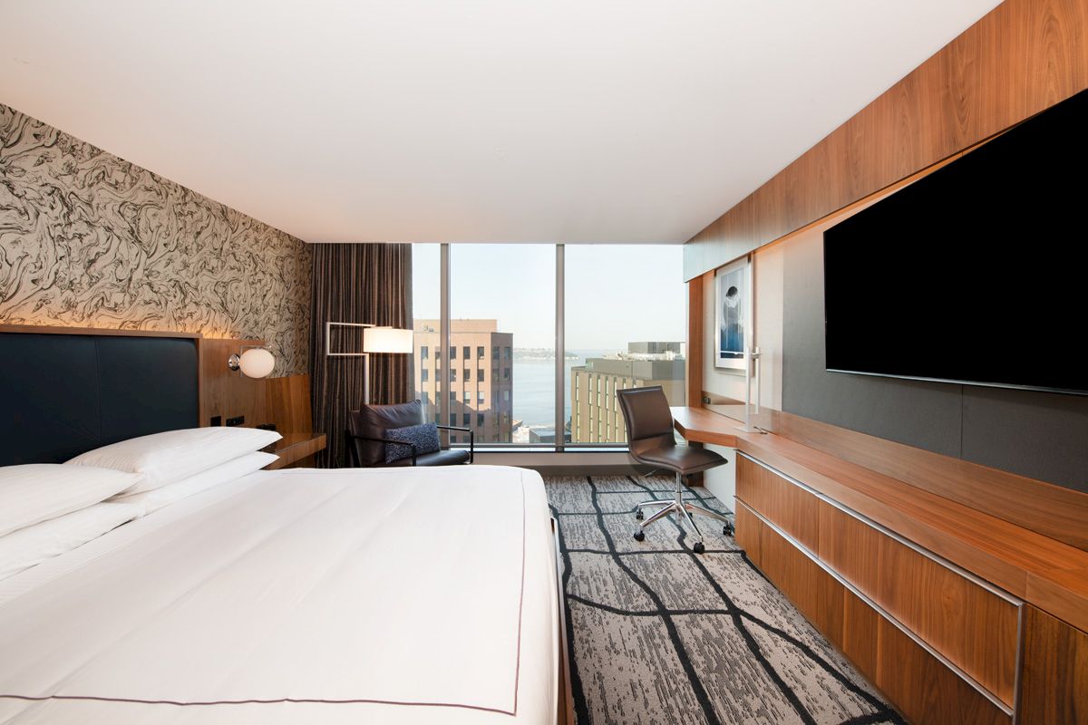 A modern hotel room with a large bed, wall art, a desk, a chair, a large TV, and a window view of city buildings and water in the distance.