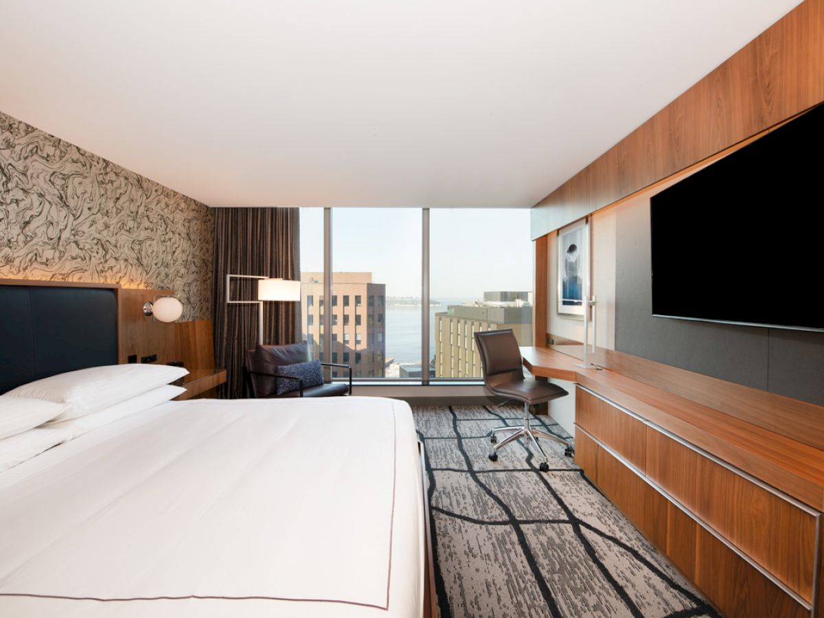 A modern hotel room with a large bed, wall art, a desk, a chair, a large TV, and a window view of city buildings and water in the distance.
