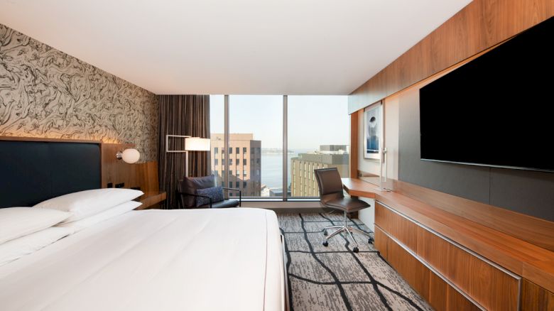 A modern hotel room with a large bed, wall art, a desk, a chair, a large TV, and a window view of city buildings and water in the distance.