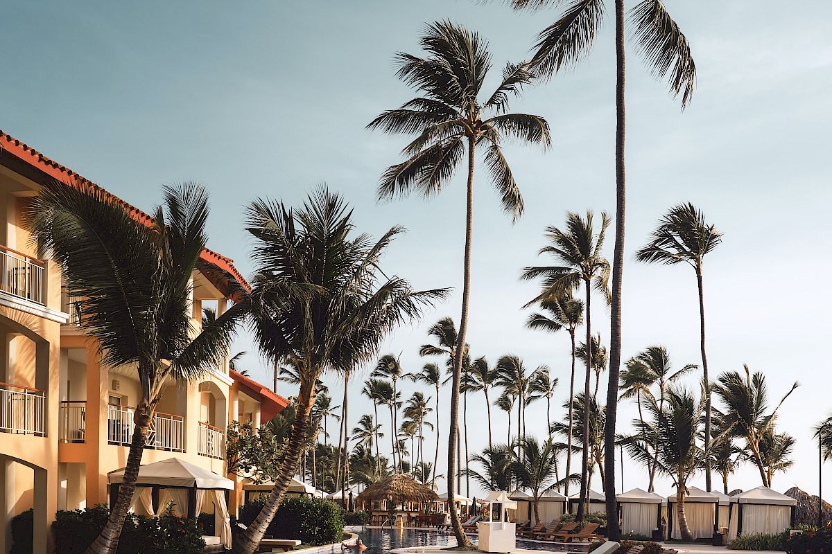 An inviting poolside scene with lounge chairs, tall palm trees, and a resort building in the background under a clear sky.