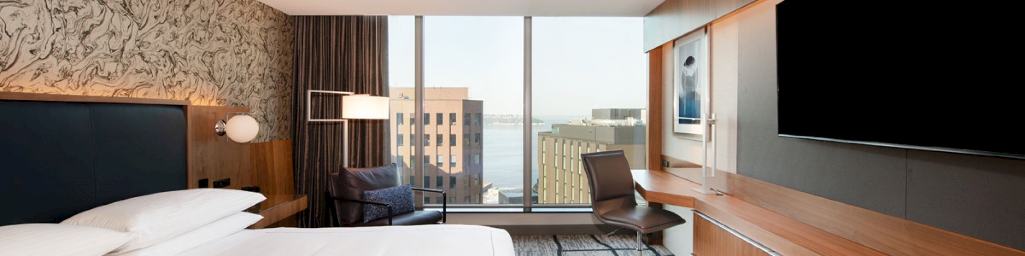 A modern hotel room with a large bed, a wall-mounted TV, a desk, and a view of city buildings through a large window.