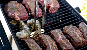 Steaks and lobster tails on a grill, seasoned and being cooked, with tongs handling the meat.
