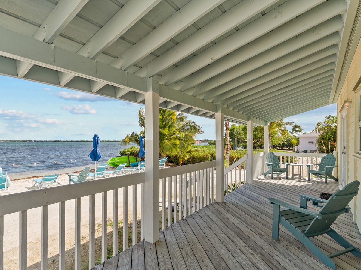 A beachfront porch with blue chairs overlooks a sandy area with more chairs and umbrellas by the water, framed by palm trees and a clear sky.