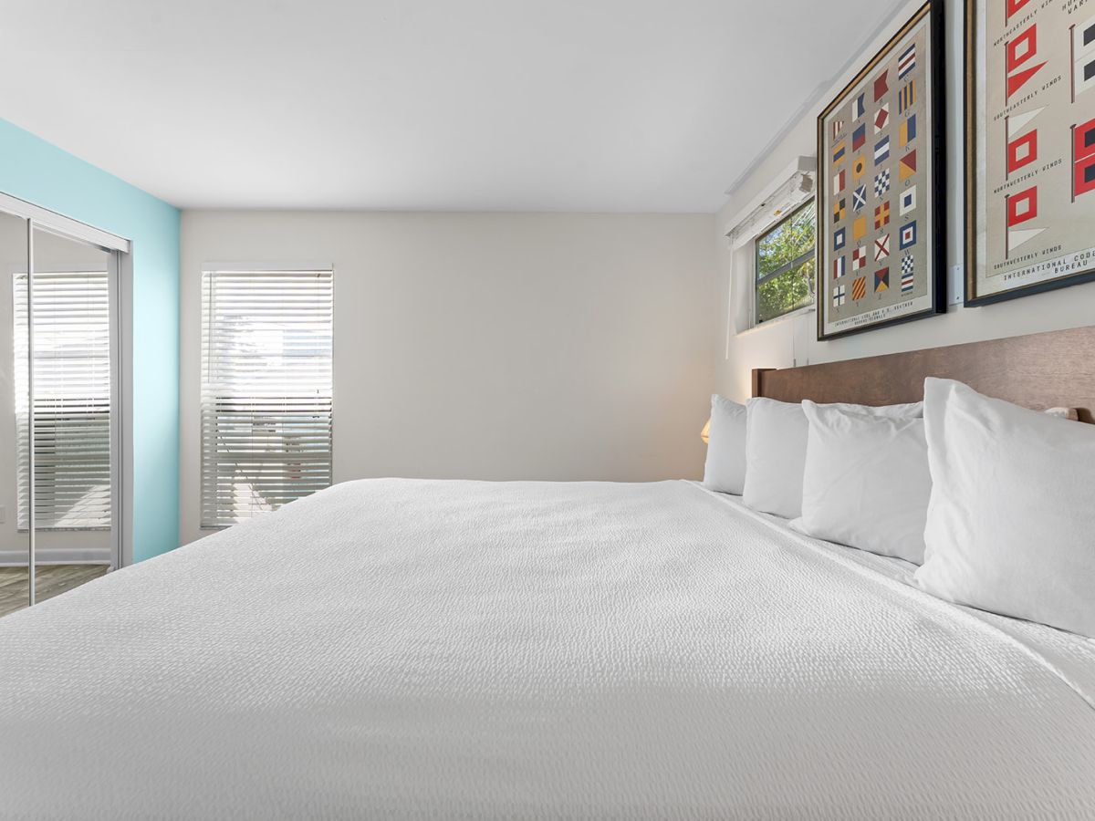 A neatly made bed in a modern bedroom, with artwork above the headboard and sunlight streaming through partially open blinds.