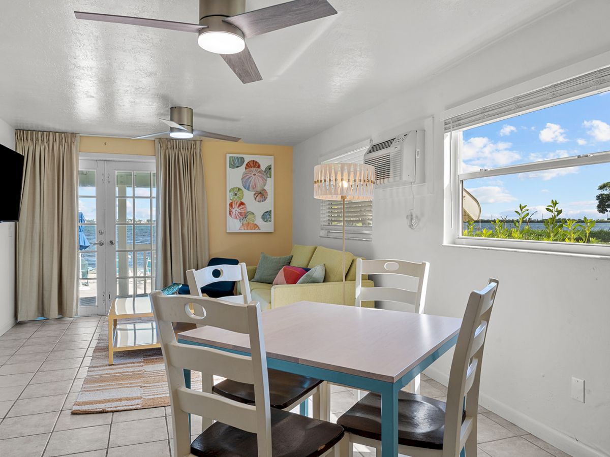 A bright, cozy coastal living-dining area with a white and blue themed table, four chairs, a beige sofa, large window view of palm trees, and balcony doors.