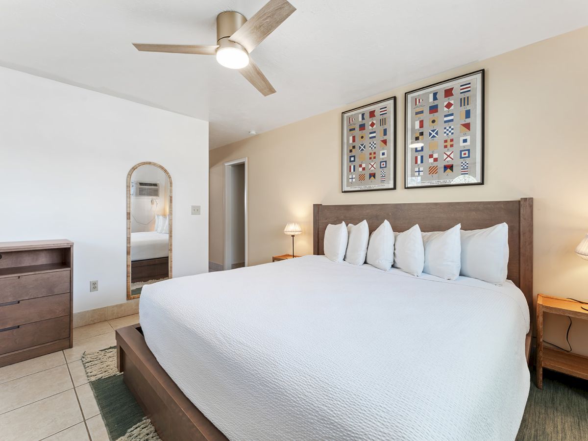 A bright hotel bedroom with a large white bed, wooden nightstands, two lamps, a dresser, a wall mirror, and framed art above the headboard.